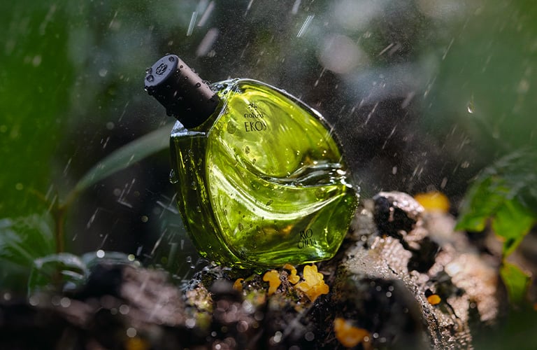 Fotografía de un frasco de perfume verde colocado sobre tierra y vegetación. El fondo muestra hojas desenfocadas y gotas de agua cayendo.